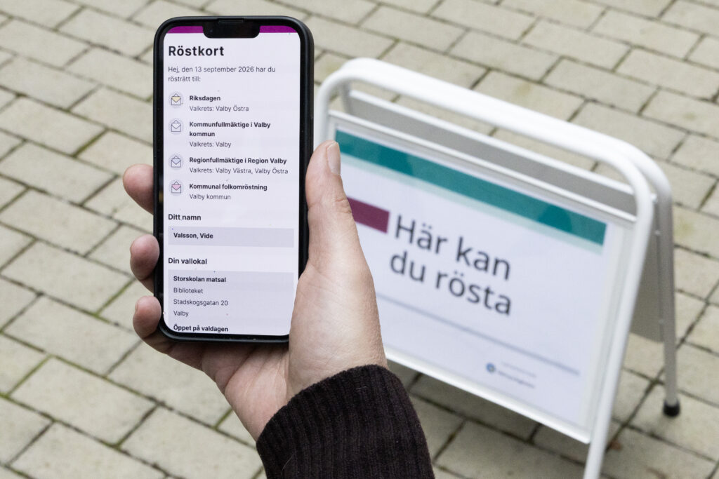 En hand som håller i ett digitalt röstkort i mobilen, framför en skylt med texten "Här kan du rösta".