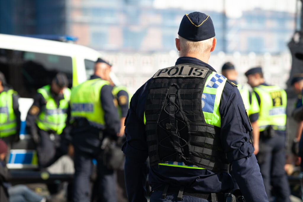 A Swedish policeman standing alone with his back turned watching his fellow police officers working in front of him.